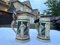 Two German-style ceramic beer steins with pewter lids, showing blue and cream color scheme and Art Nouveau female figure reliefs on white sheet background outdoors.