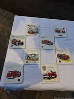 Photo displaying all eight ceramic plaques laid out on a white sheet, showing various vintage fire engine illustrations and some wear. One plaque visibly cracked.