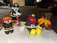 Four medium-sized plush toys displayed on white surface at night, including Dalmatian firefighter, M&M red firefighter, teddy bear in yellow raincoat, and small bear in firefighter attire.
