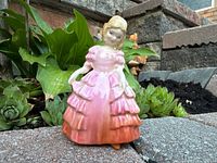 Photo of figurine standing outdoors on pavement with green foliage and stones behind it.
