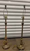 Full view of two tall brass buffet lamps with harps, placed side by side in front of a corrugated metal background. The lamps have round bases with leafy decorations and slender columns.
