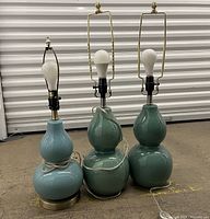 Three double gourd ceramic lamp bases arranged side by side on the floor; two green and one blue. All wired with light bulbs and no lampshades.