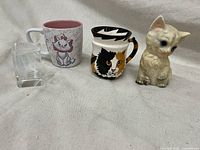 All four items seen together: two cat-themed mugs, ceramic cat figurine, and clear glass block with etched cat design.