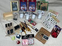 Photo of assorted craft supplies including paintbrush sets, stickers, magnets, paper punches, colored pencils, small wooden box, white candle holders, a clear measuring cup, and a framed black and white photo.