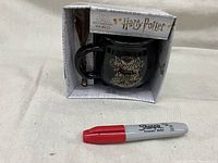 Black cauldron-shaped Harry Potter mug with a gold Hogwarts crest on the front, displayed in original packaging with wand-shaped spoon included.