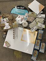 Top-down photo of assorted office and craft supplies arranged on carpet. Visible items include canvas, thick brown twine, scissors, stencil wheels, pearl beads, mini clothespins, white twine, glitter paper, envelopes, and small fasteners.