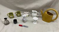 Photo showing all nine ceramic and porcelain items including pitchers, vases, small pots, and mustard-colored watering can with a Sharpie for scale.