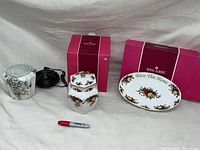 Two porcelain pieces: a floral Old Country Roses hexagonal biscuit jar with silver lid and placed beside an oval serving platter with 'Bless This Home' in floral pattern, two red boxes and a black electrical cord visible.