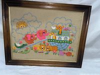 Framed vintage crewel needlepoint artwork showing colorful animals on Noah's Ark with sun and clouds embroidered in yarn on fabric.