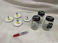Photo of three clear glass wishing jars with black lids and twine tags, three small white trinket trays each with a ceramic pineapple, one white trinket tray with a ceramic llama figure, and a red Sharpie for scale.