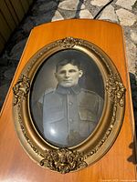Full view of the antique oval frame with decorative gold-tone accents and the black-and-white soldier portrait under convex glass, placed on a wooden surface outdoors.