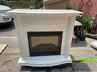 Front view of white wooden electric fireplace mantel with black heater insert installed. Classic molding details with curved bottom edge.