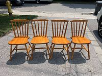 Four wooden dining chairs with spindle backs and contoured wooden seats, shown from front outdoors on asphalt.