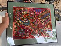 Front view of framed Mola textile art showing a rooster and dog in red, yellow, blue, and green fabric layers, with hand-stitched detail. Displayed outdoors by a bike.