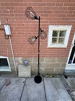 Full view of the industrial-style floor lamp with three staggered wire cage lamp heads on a matte black metal frame against a brick wall.