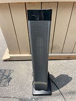 Front view of tall, black tower space heater with metal grille and base visible, set outdoors on pavement.