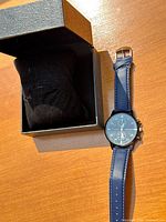 Photo showing the wristwatch placed beside an open black presentation box on a wooden surface.