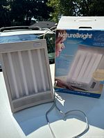 Light therapy lamp displayed outdoors on a surface, showing the front view with visible fluorescent tubes and attached power cord. Original product box standing next to the lamp with branding and product details visible.