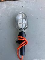 Photo of the handheld Noma work light showing the clear protective metal cage, white bulb inside, and orange power cord coiled around the black handle.