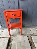 Red painted wooden side table against a dark wall showing front view with single drawer and lower shelf.