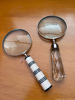 Both handheld magnifying glasses side-by-side on a wooden surface showing different decorative handles and metal rim lenses.