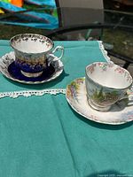 Pair of bone china teacups and saucers on teal cloth, showing one set with deep cobalt blue and gold filigree pattern and the other with floral landscape design.