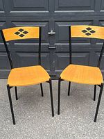 Front view of two bistro chairs with light wood veneer seats and backs, showing diamond cut-out detail on backrests and black metal legs.