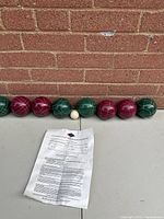 Full set of 8 bocce balls (4 green, 4 red), 1 white pallino ball in front, with printed rules sheet on table surface against brick wall.