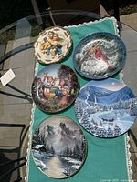 Top-down photo of all five decorative collector plates arranged on green cloth with lace trim on a glass table outdoors