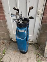 Golf clubs and blue vinyl bag standing against door on pavement, showing overall condition and completeness.