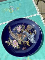 Photo of vintage Kutani porcelain plate on teal embroidered cloth, showing peacock and floral motif on a rich cobalt blue background with gold accents.
