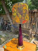 Lamp shown outdoors on wooden surface with visible purple tapered glass base, brass-tone flared base, and colorful abstract patterned fabric shade. Black cord visible coiled on top of surface.