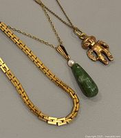 Three gold-tone necklaces displayed showing chain styles and pendants: box-link chain, chain with green drop and pearl pendant, chain with textured humanoid pendant