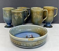 Front view showing six pedestal Irish coffee cups with gold 'Irish coffee' text and a matching bowl in front with decorative floral border and painted design inside.