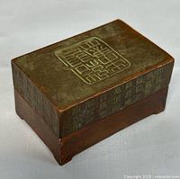 Angled photo showing top and side of the rectangular cast bronze lid with Chinese seal pattern and calligraphy, and wooden base.