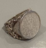 Side angle view of vintage ring showing coin top with symbols and ornate silver band with filigree details.