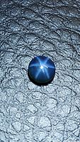 Round dark blue cabochon gemstone with a clear 6-ray star asterism under direct light on textured background.
