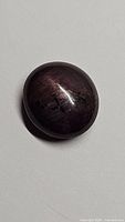 Oval cabochon cut ruby gemstone showing dark hue and surface star effect under diffuse lighting.
