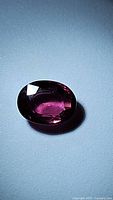 Oval cut natural rhodolite garnet gemstone, deep pink-purple color, shown from a top angle with light reflection.