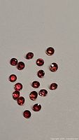 Image showing 19 small round faceted gemstones in deep red and orange hues scattered on a plain surface