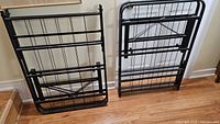 Two black metal bed frames leaning against a wall on a wooden floor, showing their sturdy metal bars and construction.