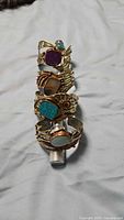 Photo of 4 gold-tone metal cuff bracelets stacked vertically and a coordinating ring, each featuring a different semi-precious stone: purple, brown, turquoise, and light blue.