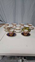 Photo showing the full set of 9 porcelain espresso demitasse cups and saucers with assorted hand-painted classical and floral designs and gold trim.