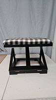 Side view of foot stool showing black and white checkered top and wooden folding legs in open position.