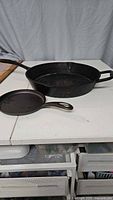 Two cast iron pans displayed on a white table, one skillet and one larger shallow pan with a spout