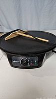 Electric crepe pan with temperature control dial and two indicator lights, black color, with power cord attached, on a white surface in front of a gray backdrop.