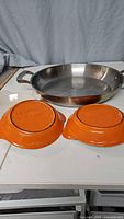 Photo showing one stainless steel paella pan with handles, placed behind two small orange Le Creuset casserole dishes upside down.