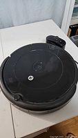Photo of black circular robot vacuum cleaner on white surface with black charging dock behind it.