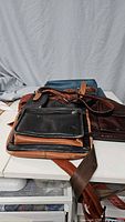 Four leather shoulder bags stacked, showing dark brown, light brown, and black colors with adjustable straps visible