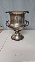 Front view of a large silver-plated metal champagne bucket displaying wide flared rim, bulbous body, pedestal base, and two curved handles. Noticeable tarnishing on surface.